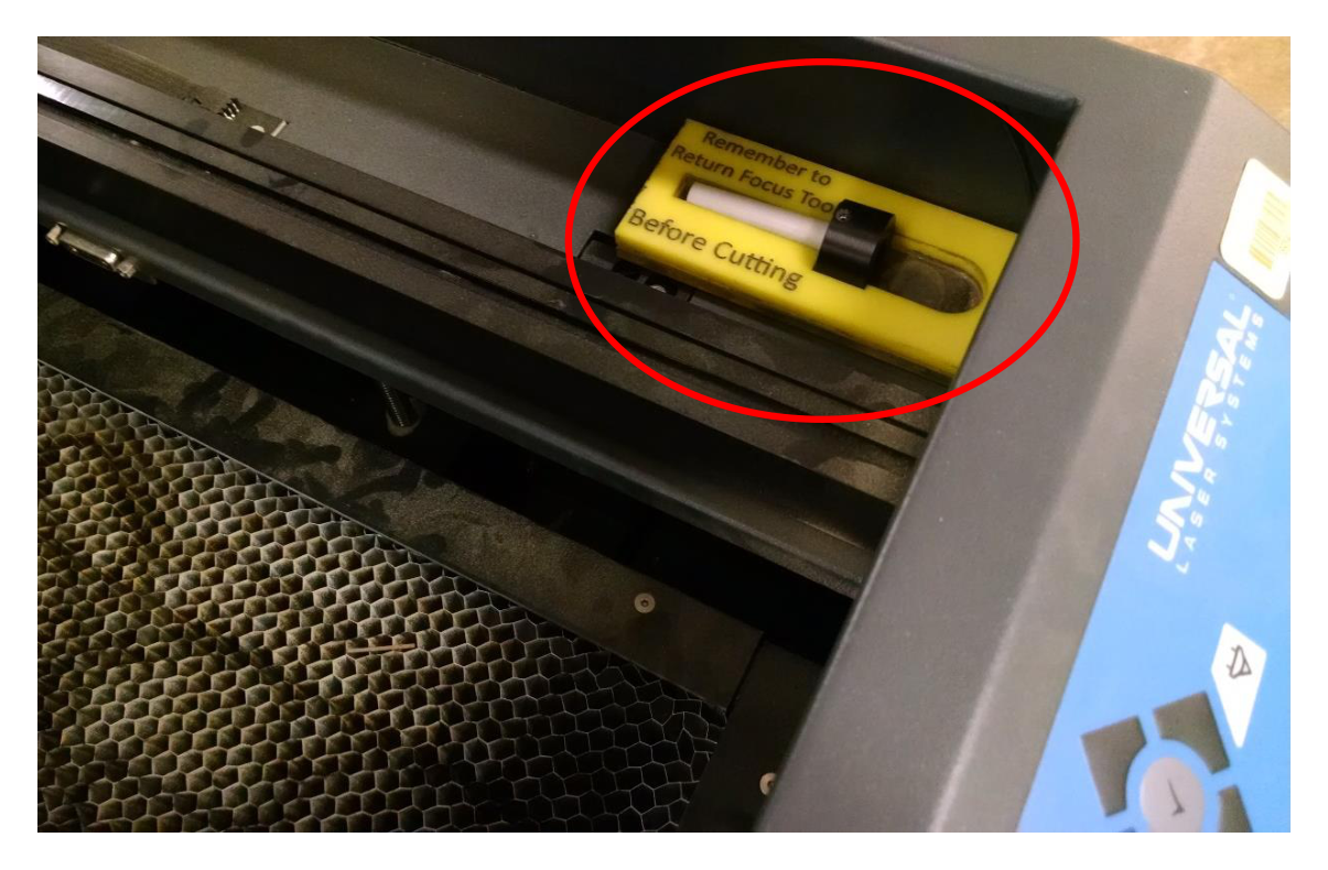 Inside of the laser cutter with the black and white plastic focus tool on the right-hand shelf circled