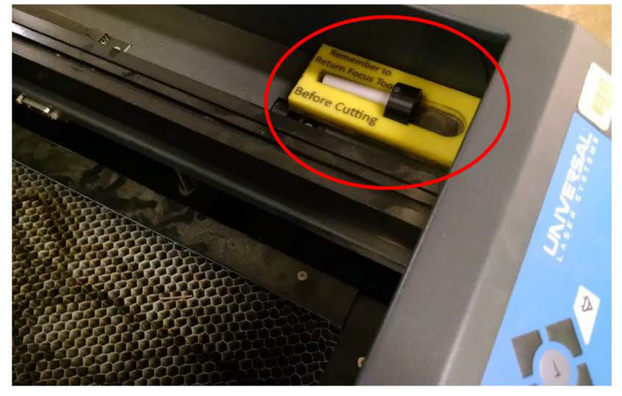 Inside of the laser cutter with the focus tool circled in red on its shelf, labeled remember to return focus tool before cutting